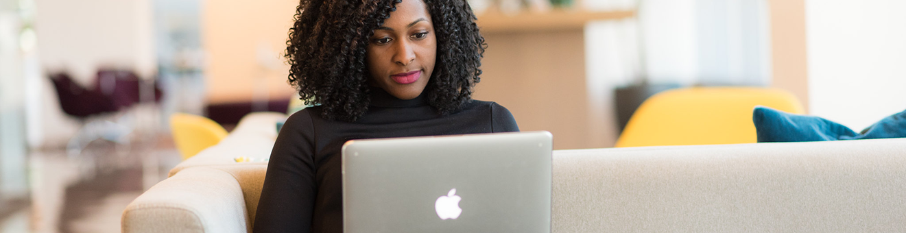 Black woman sitting on an off-white couch, working on an Apple laptop